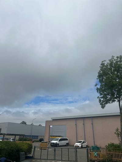 The image shows an outdoor scene with a building and a cloudy sky. The building appears to be a commercial or industrial property with a brick facade and large windows or doors. There are several vehicles parked outside, including a white car and a white van. A wooden fence and some greenery are also visible. A sign with green arrows indicating directions for people is posted on the fence.
The sky is mostly cloudy, with some patches of blue sky visible. The lighting suggests it is daytime, possibly late afternoon or early evening. The scene seems to be located in an urban or suburban area.
Visible text includes "DHL" on the side of the white van. There is also some blue text on the side of a building that appears to read "APPSTECHNIEK".