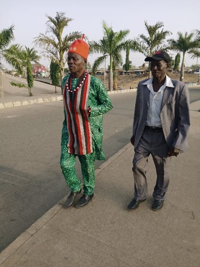 Two adult males are depicted walking outdoors on a paved surface in Mumbwa, Zambia. The individual on the left, an older male, wears a red conical hat, a multi-colored beaded necklace, and a green patterned long-sleeved tunic with matching trousers. A striped fabric in red, white, black, and grey is draped over his chest. He wears black dress shoes and looks forward and slightly left. The individual on the right, also an older male, wears a black baseball cap with a red emblem, a light grey suit jacket over a white collared shirt, dark grey trousers, and black slip-on shoes. He looks forward and slightly right. Both men are in motion, facing generally forward. The background features several palm trees of varying heights, with indistinct urban structures visible further in the distance under a clear sky. A concrete curb runs parallel to their walking path.