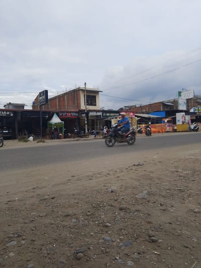 The image depicts a street scene in Gampongpaya, Indonesia, under an overcast sky. The foreground is a dusty, gravelly roadside. In the middle ground, a two-lane asphalt road runs horizontally across the frame. On the left side of the road, a row of small shops with corrugated metal roofs are visible. One shop has a black sign with the text "A AMIRSTYLE". A dark-colored car is parked in front of this establishment. Further down the row, a tent-like structure is set up, with several people standing around it. A motorcycle is parked nearby. On the right side of the road, more shops are present, including one with a sign that reads "WM BOUTIQUE". Several motorcycles are parked or being ridden on this side of the road. One man is riding a motorcycle in the foreground, moving from left to right, appearing slightly blurred due to motion. Behind the shops on the right, there are buildings under construction with exposed brickwork and concrete. Several power lines are strung overhead. The overall impression is of a bustling, informal commercial area.