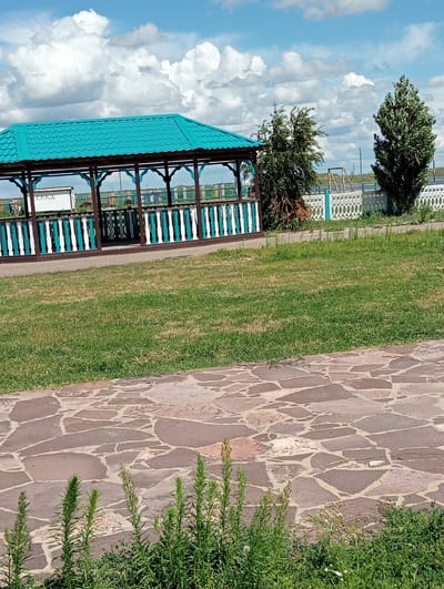 A sunlit, outdoor scene depicts a gazebo with a turquoise roof, supported by dark brown posts and enclosed by a decorative fence with white and teal vertical slats. White text, reading "КЛАД," is visible on a small white structure behind the gazebo. Adjacent to the gazebo, a paved walkway made of irregularly shaped reddish-brown stones leads the eye towards a grassy area with sparse vegetation in the foreground and two trees. In the background, a white, decorative fence with circular motifs runs horizontally, with a soccer goal visible further back. The sky above is a bright blue, dotted with fluffy white clouds. The location is identified as Zhanyspay, Kazakhstan. There are no people clearly visible in the frame.