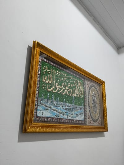A framed depiction hangs on a pale gray wall. The art presents a detailed image of a mosque with numerous minarets and architectural details. Above the mosque, calligraphic Arabic script is prominently displayed in golden hues. A clock is situated on the right side of the framed piece, adding a functional element to the artistic representation. A golden frame surrounds the entire artwork. The lighting accentuates the details and adds depth to the composition.