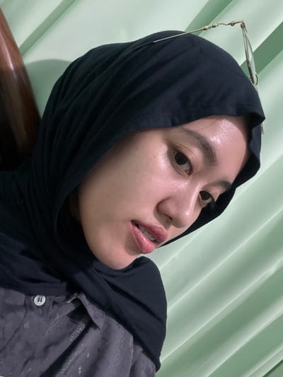 An angled, close-up portrait in Patrang, Indonesia, features a person, likely a young woman, wearing a black hijab and a dark grey button-up shirt with a subtle, lighter-toned pattern. A pair of thin, gold-framed eyeglasses rests atop her hijab. Her face, seen from the chin up, exhibits light skin tones with visible highlights on the forehead and nose. Dental braces are visible on her upper teeth. Her dark eyes are directed downwards and slightly to the right. The background consists of a light green, vertically ribbed surface, possibly a wall or curtain. A dark, smooth, vertical object, potentially wood, is partially visible along the far left edge of the frame. The subject appears to be still, posing for the photograph.