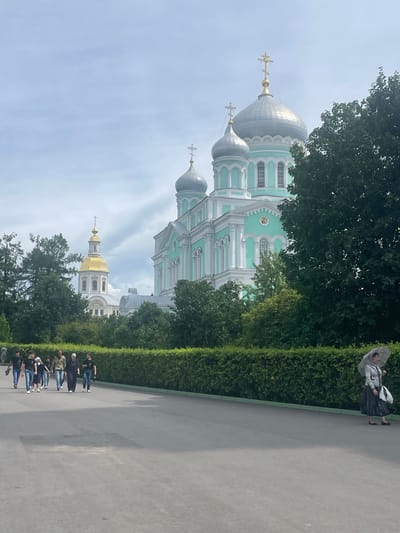 The image shows a group of people walking along a paved path in front of a large, ornate church with multiple domes in Diveyevo, Russia. The church is predominantly light teal and white, with silver and gold domes. A taller, separate bell tower with a golden dome is visible to the left of the main church. Lush green trees and hedges line the path. In the foreground, a group of young people, appearing to be teenagers and young adults, walk together, some looking at their phones, others talking. Further down the path, a woman walks alone, holding an umbrella. The sky is overcast, with scattered clouds.