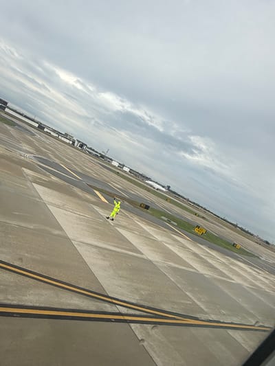 The image depicts a view from an elevated position, likely an aircraft window, overlooking an airport tarmac under an overcast sky. The tarmac is concrete and marked with yellow and black lines. Small patches of grass are visible between the concrete sections. Yellow signs with black lettering are interspersed throughout the tarmac. A person wearing a high-visibility yellow suit is visible on the tarmac. The general location is the airport in San Antonio, United States of America.