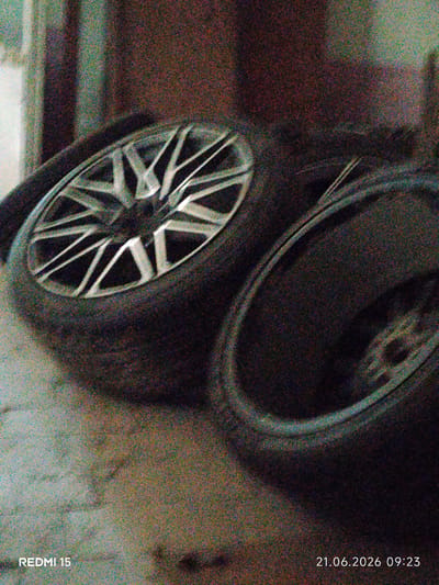 The image depicts two car tires, one with a visible silver alloy rim. The tires are positioned on a rough, textured surface, possibly concrete or stone, in what appears to be an outdoor or semi-outdoor environment. The background is dark and indistinct, with vertical lines suggesting a wall or structure. A watermark in the lower left corner reads "REDMI 15", and in the lower right corner, a date and time stamp indicates "21.06.2026 09:23". There is significant image noise, which affects the clarity and detail of the scene. The location of Yovon, Tajikistan is not discernible from the visual information present in the image.