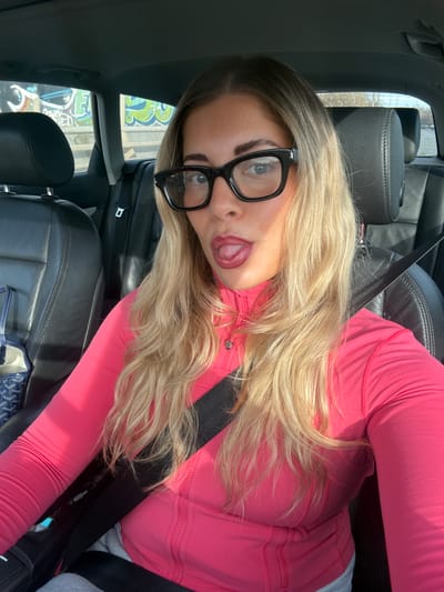The scene depicts a light-skinned female with long, wavy blonde hair, visible darker roots, and brown eyebrows, positioned in the driver's seat of a vehicle, purportedly in Marcq-en-Barœul, France. She wears large, black-framed rectangular eyeglasses and red lipstick. Her expression is characterized by a slightly extended tongue. She is dressed in a bright pink, long-sleeved athletic top with a partial zip closure, featuring a small, circular logo consistent with Lululemon branding on the zipper pull. A black seatbelt crosses diagonally over her torso. Grey fabric, consistent with sweatpants, is visible below the seatbelt. On the unoccupied front passenger seat, a blue and white patterned tote bag is partially visible. The car interior features dark leather seats in both the front passenger and rear sections. Through the rear passenger window, a concrete wall adorned with colorful graffiti in shades of blue, green, and white is visible, indicating an urban or roadside environment. Strong natural light illuminates the subject from the right (passenger) side. The female is looking directly into the camera, capturing a selfie.