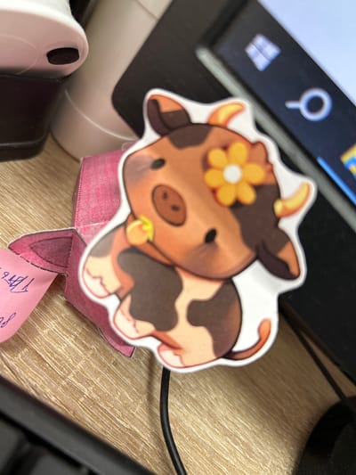 This is a close-up, eye-level shot of a decorative paper cutout of a cartoon cow sitting on a wooden desk. The cow is brown and white with a yellow flower on its head and a small green ring around its neck. The cutout is attached to a pink, folded paper base. To the left of the cutout, a portion of a white and black electronic device is visible, likely part of a computer or printer. In the background, a computer screen displays a search icon and a blurred image, suggesting an office or work environment. A black cable snakes across the desk behind the cow. The lighting appears to be artificial, indicating an indoor setting during the day. No specific time of day or weather conditions are discernible. The location context provided is Пекалінскі сельскі Савет, Belarus, but there are no visual cues within the image to confirm this specific location. A small piece of pink paper with handwritten blue script is visible sticking out from under the cow's base, but the text is illegible.