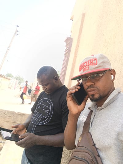 The scene depicts an outdoor urban environment in Zango, Nigeria, under bright daylight. In the immediate foreground, two adult males are visible. The man on the right, with a dark complexion, wears a grey polo shirt, a light-colored baseball cap with "CHICAGO CHI" lettering in red, rectangular glasses, and white wireless earbuds. He holds a black smartphone to his right ear, actively engaged in a call, and looks towards the viewer. A brown fanny pack or sling bag is visible across his chest. To his left, another adult male, also dark-complexioned with a shaved head, wears a black t-shirt with a white stylized circular graphic (resembling "FF") and blue jeans. He is looking down and manipulating a black smartphone or small tablet with both hands.

In the midground and background, an unpaved street or path extends, lined with various light-colored, single-story buildings. Several other individuals are present further down the path, including one person in a red shirt walking left and another in a red shirt standing. A utility pole with overhead wires is visible on the left side of the frame. To the right of the foreground subjects, a light beige building with a decorative column is partially visible. A low concrete structure runs horizontally across the midground. The sky is largely overexposed, indicating strong sunlight.