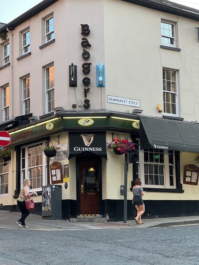 The image shows the exterior of a pub named "Badgers" at the corner of Newmarket Street in the city of Derry/Londonderry, United Kingdom. The pub has a distinctive green awning and signage, as well as a Guinness advertisement. Two women are visible: one in the foreground walking left to right with a red bag, and another in the background walking away from the camera. The street is paved, and a traffic light is visible. The building is old and has multiple windows. The overall impression is a typical street scene in a historic city.