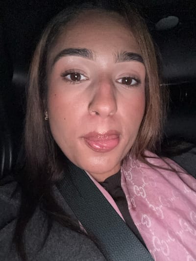 A woman's face is centered in the frame. She has brown eyes, thick eyebrows, and a nose ring. Her lips are glossy, and she wears blush. A pink Gucci scarf is draped around her neck, and a seatbelt crosses her chest. The background is dark, suggesting she is in a car.