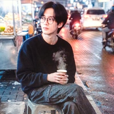 In the city of Cigadung, Indonesia, a young East Asian man with dark hair and glasses sits on a light-colored stool. He is wearing a dark crew neck sweatshirt and grey cargo pants. In his hands, he holds a white paper cup from which steam rises. The background is blurred but appears to be a street scene at dusk or night, with streetlights and traffic, including a white car and motorcycles. To the left, a food stall with various items is visible. The ground is wet, suggesting recent rain.