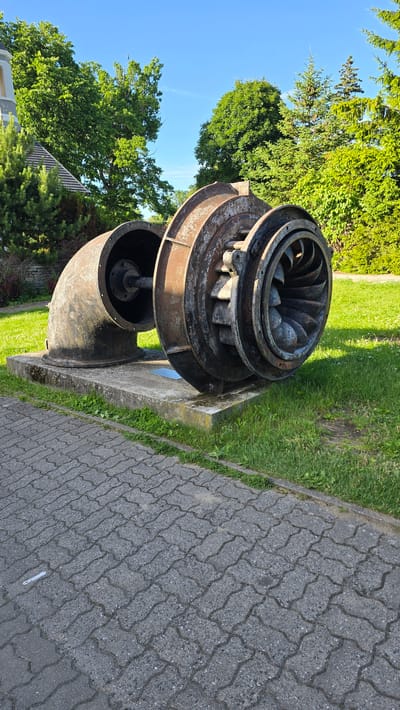This image shows a large, rusted turbine wheel and casing, likely part of a hydro-electric power plant. The turbine is made of dark metal with visible signs of age and weathering, including rust and some wear on the blades. It is mounted on a concrete base, which is partially covered by grass. To the right of the turbine, there is a paved walkway with interlocking grey pavers. The walkway is bordered by a strip of green grass. In the background, lush green trees and a bright blue sky are visible. The overall scene suggests an outdoor setting, possibly a park or an industrial site repurposed for public display. The lighting indicates it is daytime, with clear weather. No people are visible in the image. The location context provided is Keila-Joa alevik, Estonia, which is known for its historical water mill and waterfall, making the presence of a turbine plausible. There is no visible text in the image.