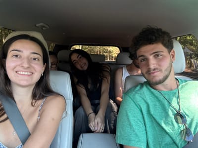 The image displays five individuals inside a vehicle, likely an SUV or minivan, in Otego, United States. A young woman with long dark hair, wearing a light-colored patterned top and a seatbelt, smiles directly at the camera from the front passenger-side seat. To her right, a young man with dark curly hair and a beard, wearing a green t-shirt and a dark beaded necklace with sunglasses, looks towards the camera with a neutral expression. Behind the woman on the left, a second young woman with dark hair, wearing dark clothing and rings, is partially visible, also looking towards the camera with a slight smile. Behind the man on the right, a person wearing a white tank top is partially visible. Further back, mostly obscured by the front occupants, a fifth person wearing glasses is dimly visible. The vehicle's interior features light-colored seats and a dark headliner. Through the windows, a sunlit wooded environment with dense green trees and a distant white tent-like structure is visible.