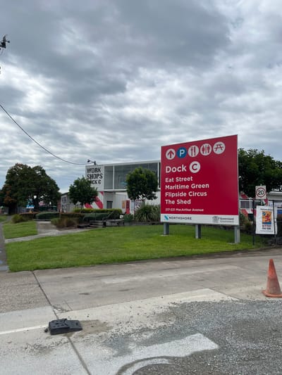 An outdoor scene in Hamilton, Australia, features a large red directional sign prominently on the right, under an overcast, cloudy sky. The sign displays white icons for parking, food services, restrooms, and a picnic area, each with an upward arrow. Text on the sign indicates destinations including "Dock C," "Eat Street," "Maritime Green," "Flipside Circus," and "The Shed." It also provides the address "217-221 MacArthur Avenue" and features logos for "Queensland Government" and "NORTHSHORE," identifying the precinct as Northshore Hamilton.

The ground consists of a paved road or pathway, bordered by neatly maintained green grass. In the background, to the left of the main sign, is a white sign on a building with "WORK SHOPS" and "DOCK C" printed on it. This area appears to be an industrial or commercial zone, possibly featuring structures made from converted shipping containers. Further along the pavement to the right, an orange traffic cone is visible, along with a "20" speed limit sign and a small, black utility box on the ground. Another partially visible sign in the background contains the text "BUTTERY". The scene depicts a quiet daytime setting, with no people or specific activities taking place, focusing on infrastructure and wayfinding within the Northshore Hamilton precinct.