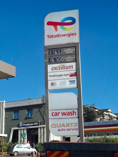 The image shows a TotalEnergies gas station located at coordinates -1.227, 36.664 in Gitaru, Kenya. The gas station sign displays the prices for petrol and diesel: 176.50 for petrol and 167.00 for diesel. The sign also advertises "excellium" fuel and TotalEnergies Card payment options. Below the price display is a sign advertising a car wash and "QUARTZ auto service." The station has a small, single-story building with a Tropicana Cafe sign. A silver car is parked in front of the building, and a person is walking away from the car toward the building. The sky is blue and clear, with no clouds visible.