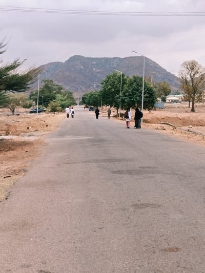 The image displays a paved road stretching into the distance in Rafin Pa, Nigeria, under a cloudy sky. A large, rugged mountain range dominates the background. The road's surface is gray asphalt, exhibiting minor wear. Along the left side of the road, dry, reddish-brown earth with sparse green and dry vegetation is visible, including several trees, one appearing leafless. A dark-colored vehicle is parked on the left side in the mid-ground. On the right side, a line of trees with dense green foliage runs parallel to the road, and a tall, lighter-colored tree with bare branches stands further back. In the far distance on the right, low-rise buildings with light-colored roofs are discernible. Multiple white-poled streetlights are positioned along both sides of the road.
Ten individuals are present on the road, generally moving away from the camera. In the mid-ground on the left, two individuals wearing white long-sleeved tops and dark lower garments are walking side-by-side, one carrying a white bag. Further down the road on the left, another person in dark attire is walking. On the right side of the road, two individuals are standing facing away from the camera; one wears a dark top and light pants, and the other wears a white top, dark lower garment, and reddish footwear, with a dark bag on their back. Further back on the right, a person in dark clothing walks, followed by another individual in light brown attire.