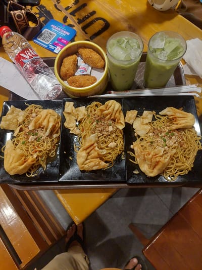 This overhead shot captures a meal at what appears to be a casual eatery. The table is a bright yellow, with a wooden table leg visible on the right. On the table, there are three black square plates, each holding a serving of noodles topped with fried wontons and what looks like minced meat and crispy fried onions. Beside the plates, a bamboo steamer basket contains four breaded, fried items, possibly spring rolls or chicken nuggets, with small packets of sauce. Two tall glasses filled with a green, icy beverage, likely matcha or avocado juice, stand to the left. A bottle of Vit water and a receipt are also present.
A blue QR code sign with Indonesian text, "PESAN DISINI TANPA ANTRI" (Order Here Without Queueing), suggests a self-service ordering system. The overall scene implies a relaxed mealtime, perhaps lunch or dinner. The lighting is bright, suggesting it could be daytime, and there are no obvious cues about the weather. The presence of the QR code and the type of food points towards a modern, casual dining establishment in Indonesia, potentially Pematang Siantar, as indicated by the water bottle brand. A person's feet are visible in the lower right corner, indicating someone is seated at the table. The atmosphere seems laid-back and inviting, focused on enjoying the food and drinks.