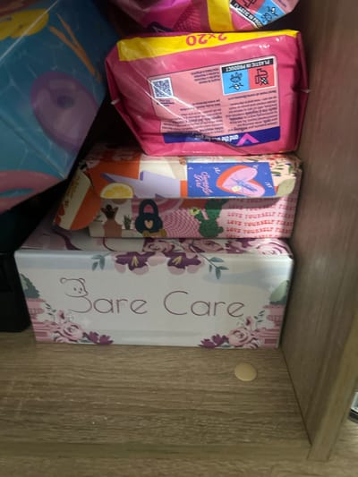 The image shows a stack of items on a wooden shelf. The top item is a pink package with a yellow stripe, labeled "2x20". Below it is a patterned box with the words "Sweety Girl" and a heart graphic. The third item is a box with repeated text "LOVE YOURSELF FIRST" and graphics of cacti and hearts. The bottom item is a white box with floral patterns and the text "Bare Care" and a cartoon bear graphic. The background appears to be a wooden shelf or cabinet. There are no people visible in the image. The overall impression is a collection of personal care or feminine hygiene products. The image does not provide any information about the city of Perushtitsa, Bulgaria, or any specific location within it.