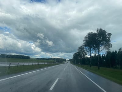 The image shows a highway in Ракаўскі сельскі Савет, Belarus. The road is wet and reflects the overcast sky, which is filled with dramatic, heavy clouds with patches of blue sky peeking through. Trees line the right side of the road, with a dense forest in the distance on the left. The highway has white lane markings and a guardrail. There are a few cars visible in the distance on the road, and some poles with red tops are positioned along the side of the road. The overall impression is of a slightly gloomy, but scenic, rural landscape.