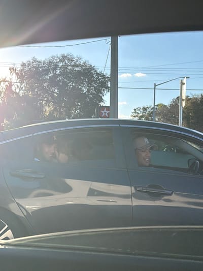 The image depicts a dark-colored sedan, likely gray or black, with two visible male occupants, captured from an adjacent vehicle. The scene is set in Gainesville, United States, near a Texaco gas station, identifiable by its red star logo visible in the background.
The driver, an adult male of apparent East Asian descent, wears a light-colored New York Yankees baseball cap and eyeglasses. He is smiling and looking towards the right side of the frame. In the rear seat, directly behind the driver, a second adult male wears a dark New York Yankees baseball cap. He is smiling and looking directly at the camera, with his right hand raised near his face in an ambiguous gesture.
The background features a large, leafy tree with Spanish moss, consistent with the Florida climate. Utility poles, power lines, and a street light pole are also visible against a clear blue sky with sparse clouds. The lighting indicates a low sun angle, with prominent lens flare emanating from the left side of the frame. Both visible individuals exhibit positive facial expressions.