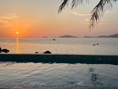 An outdoor scene captured at sunset in Ban Ko Li Pe, Thailand, features an infinity pool foregrounding a calm sea. The pool's edge, constructed with small dark mosaic tiles, occupies the lower third of the frame, its water reflecting the sky and exhibiting gentle ripples. The sun, a prominent orange disc, is positioned just above the horizon on the left, casting a shimmering path of light across the water. Several large, dark rocks are partially submerged in the sea near the pool's left side. In the midground, two individuals in kayaks or small boats are visible on the water towards the right, while a traditional longtail boat moves horizontally further to the right. Distant islands or landmasses are silhouetted against the horizon. The sky displays a gradient of warm orange, pink, and light blue hues, with sparse, wispy clouds. Dark palm fronds intrude into the upper right corner of the frame.