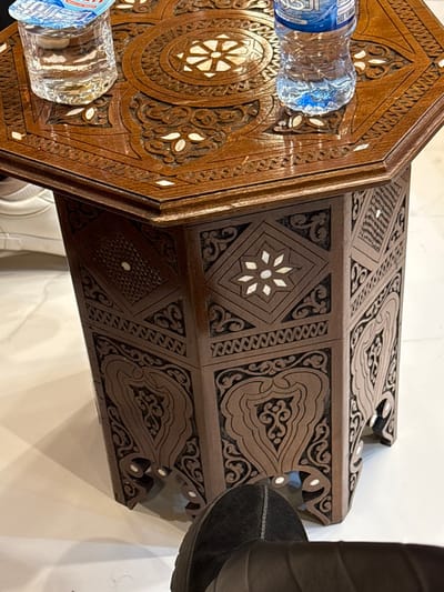 A detailed, ornately carved, brown wooden table sits on a white surface. Its octagonal top holds two plastic water bottles. The table's side features repeating geometric and curvilinear patterns, accented with light-colored inlays. A black suede shoe is partially visible near the edge of the table.