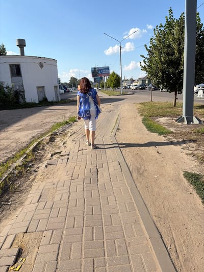 The image depicts a sunny day on a paved pedestrian walkway in Tambov, Russia. A woman with dark hair, wearing a blue and white patterned blouse and white capri pants, walks away from the camera towards the right side of the frame. She carries a white strap bag over her left shoulder. The walkway is made of grey interlocking pavers, some of which appear uneven or damaged. Adjacent to the walkway is a sandy area with patches of dry grass. In the background, a white cylindrical building with two doors and a window is visible on the left, with graffiti on its walls. Further back, a street with several cars driving on it is seen. A large billboard advertising a lottery is also present. On the right side of the frame, a tall grey pole, likely a lamppost, stands next to a leafy green tree. The sky is clear blue with a few white clouds. The overall scene suggests an urban or suburban environment.