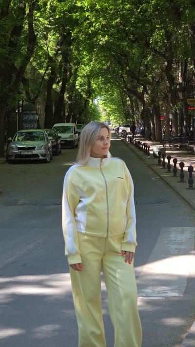 A full-body portrait captures a woman standing on a paved street lined with trees, likely in Subotica, Serbia. The woman has blonde hair and is wearing a light yellow tracksuit with white stripes along the arms and sides of the jacket and pants. She is facing slightly to her right, looking upwards with a neutral expression. Her right hand, adorned with orange nail polish, is visible near her waist, and her left hand, also with orange polish, is near her left thigh.
The setting is an urban or suburban street, characterized by a dense canopy of green trees creating an avenue-like effect. Strong sunlight casts distinct shadows on the ground, indicating a clear, sunny daytime. A row of black bollards is visible on the street beside the woman. To her left, several cars are parked, including a silver Skoda with the Serbian license plate "SU 079-IA" (indicating Subotica). Other cars are partially visible further down the street. The overall scene suggests a pleasant day for an outdoor photoshoot. The image itself is oriented vertically, implying it has been rotated from its original landscape or portrait capture.