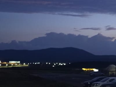 The image captures a dusk or nighttime scene in the vicinity of Баянауыл ауылдық округі, Kazakhstan. In the mid-ground, a large illuminated sign spelling out "БАЯНАУЫЛ" is visible, flanked by several streetlights. Beyond the sign, more lights indicate structures or an area with activity. The background is dominated by the dark silhouette of a mountain range under a deep blue sky with some wispy clouds. In the foreground, several cars are parked. One car, a white sedan, is in the immediate foreground on the right. To the left of it, a darker sedan is partially visible. Further to the left and slightly behind, a row of cars is parked, including what appears to be a white SUV and other darker vehicles. A white tent-like structure or yurt is also visible to the right of the illuminated sign. The overall atmosphere is dark and somewhat hazy, typical of low-light photography.