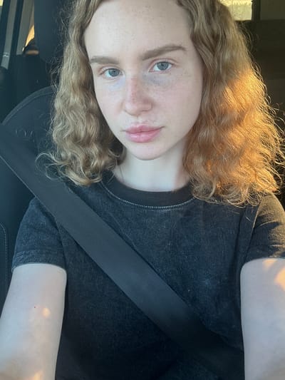 A young person with light skin, freckles, and wavy blonde hair is sitting in the passenger seat of a car. They are wearing a dark gray t-shirt and a black seatbelt. The sun is shining from the right side of the frame, illuminating their hair and left arm. The background of the image is dark and out of focus, suggesting the interior of a vehicle. There is no identifiable location in the city of Vasilyevo, Russia.