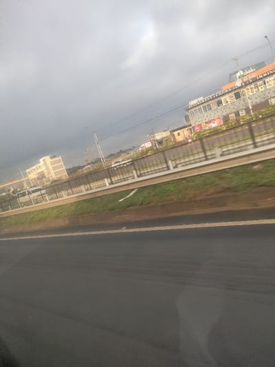The image displays an urban roadside scene in Theta ward, Kenya, under an overcast sky. The foreground features a dark asphalt road with a faint yellow line delineating a lane. A grassy median separates this road from a concrete barrier topped with a metal guardrail. Beyond the barrier, multiple vehicles, including a white van or SUV, are visible on an adjacent thoroughfare or parking area. In the background, a two-story commercial building on the right features signage reading "RESTAURANT | CHOMA ZONE" and "TASTIES IMMING," with an additional sign for "Shaki" above. To the left, a large, light-colored multi-story building is visible. Overhead power lines traverse the frame. No individuals are discernible, and no specific actions or interactions are depicted.