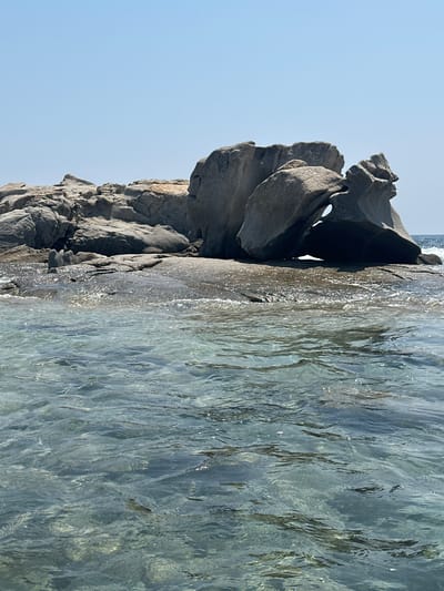 The image shows a close-up view of a rock formation jutting out of the clear, turquoise water near the coast of Castiadas, Italy. The rocks are large and weathered, with a distinctive archway formed by two boulders. The water is calm and shallow, allowing the sandy bottom to be visible. The sky above is clear and blue, indicating a sunny day. The scene is tranquil and natural, with no signs of human activity or man-made structures.