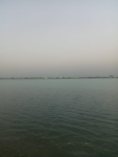 The image shows a body of water, likely the Arabian Gulf, with a distant shoreline visible under a hazy or overcast sky. The shoreline is dotted with what appear to be buildings and structures, suggesting a populated area. The water is calm with gentle ripples. The foreground is the water's edge, where the water appears darker.
Based on the visible features, it is likely that this image was taken in Al Khobar, Saudi Arabia, given its location along the Arabian Gulf and the presence of buildings along the shoreline.