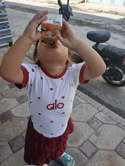 A young child is drinking from a juice box while standing on a patterned tiled patio. The child is wearing a white t-shirt with red hearts and the word "alo" on it, and a red skirt. In the background, a black motorcycle is parked. The scene appears to be outdoors, possibly in a residential area or public space within Gulistan, Uzbekistan. The lighting suggests it is daytime.