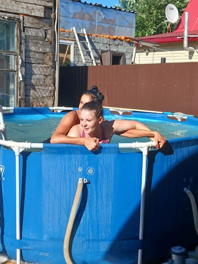 In the city of Chik, Russia, two young women are enjoying a sunny day in a blue inflatable swimming pool. The pool is set up outdoors, with a rustic wooden building and a brown metal fence visible in the background. One woman, in a pink bikini, is leaning on the edge of the pool, her arms resting on it, while the other woman, in a dark swimsuit, embraces her from behind. The water is clear, and the sun casts shadows on the pool's exterior. The scene appears to be a casual, outdoor leisure activity in a residential area.