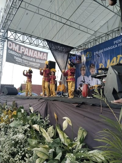 The image shows a stage in Sambirejo, Indonesia. Three young girls in traditional maroon and gold costumes are performing a dance on the stage. Behind them, a backdrop indicates the event is sponsored by "OM. PANAMA Dangdut Jadulnya Sragen" and "PIMPMAS Kantit Darwanto, SH." There is also a banner celebrating an anniversary. Additional musicians and equipment are visible behind the dancers. The stage is decorated with flowers and foliage.