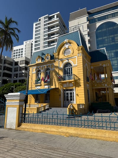 This is a vertical, eye-level shot of a bright yellow building with blue trim, set against a clear blue sky. The building appears to be historic, with ornate architectural details like decorative window surrounds and balconies. Flags are visible on the building's facade, including what appears to be the flag of the United States, along with other international flags. String lights are hung across the balconies, suggesting it may be an establishment open in the evening.

The building is situated in a dense urban environment, with modern, white high-rise buildings visible behind and to the side of it. A palm tree frond extends into the frame from the upper right, indicating a tropical or subtropical climate. The foreground shows a wooden walkway or pier. The lighting suggests it is daytime, likely mid-morning or afternoon, with strong sunlight casting shadows. The scene evokes a lively atmosphere, possibly a commercial or tourist area in Santa Marta, Colombia. There is no visible text in the image.