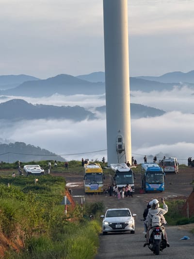 This is a vertical, eye-level shot taken in Phát Chi, Vietnam, capturing a scenic view dominated by a large white wind turbine. The turbine is positioned on a hilltop, with a vast expanse of fog-filled valleys and layered mountains stretching into the distance under a hazy sky.

Several vehicles are present, including a white car in the foreground, a motorcycle with two passengers, and multiple tour buses parked on a dirt road. A white, futuristic-looking car is tipped on its side further up the hill.

A crowd of people are gathered around the base of the wind turbine and near the parked buses, appearing to be tourists. Some are taking photos, while others are engaged in conversation. The surrounding landscape is a mix of green grass, vegetation, and reddish-brown earth. A traffic signpost with red and white markings is visible on the side of the road. The overall scene suggests a popular tourist destination with a dramatic natural backdrop.