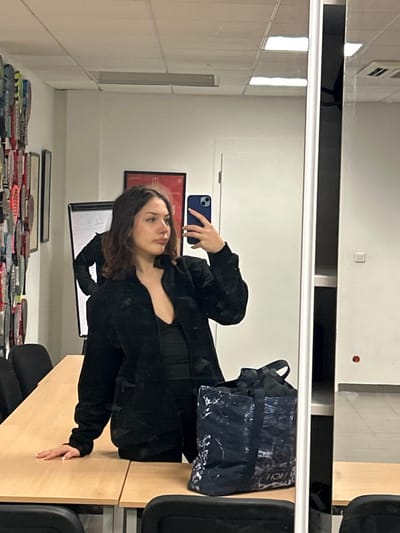 A mirror selfie of a woman with wavy brown hair wearing a black jacket is the focal point. She holds a blue phone. The background includes a white wall with vents, a red-framed picture, and a whiteboard. Below, a stack of red and blue objects is present. A desk with a blue bag and black chairs occupies the lower right corner of the image.
