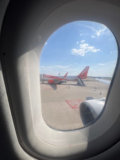 The image, taken from an airplane window, shows an EasyJet airplane on the tarmac. The plane is predominantly white with a distinctive orange tail fin featuring the "easyJet" logo. A jet bridge is connected to the aircraft, suggesting boarding or deplaning is in progress. The plane's engines are visible, with one appearing larger in the foreground.

The setting is an airport tarmac under a clear blue sky with scattered white clouds. The ground is paved concrete, marked with red and white lines, possibly indicating parking or taxiing areas. An orange traffic cone is visible on the tarmac. The time of day appears to be daytime, with bright sunlight casting shadows.

The view is framed by the rounded interior of the airplane window, with the greyish plastic frame dominating the foreground. The overall impression is one of travel and anticipation. No people are clearly visible in the image. The location is identified as Paray-Vieille-Poste, France.