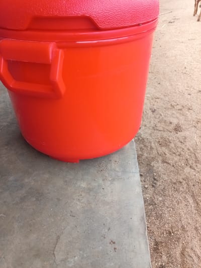 The image shows the side of a large, bright red plastic cooler. The cooler has a textured lid and a sturdy handle attached to the side. It is positioned on a flat, grey concrete surface. The background consists of sandy, dry ground, with a few small pebbles and some debris. In the far upper right corner, the leg of an animal, likely a goat or a cow, is visible, suggesting an outdoor, potentially rural setting. The overall scene appears to be a still life, focusing on the cooler. The location in Zinna, Nigeria, is not visually indicated in the image itself but is provided as context for the scene.