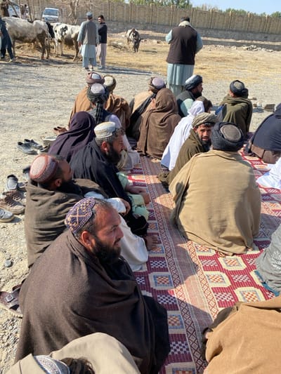 A group of men in traditional clothing and hats are praying on a woven mat outdoors. They are lying prostrate, some covered in blankets or shawls. In the background, other men stand near cows, a vehicle, and a dirt road. A low wall is visible in the distance. The overall scene suggests a religious observance in a rural setting.