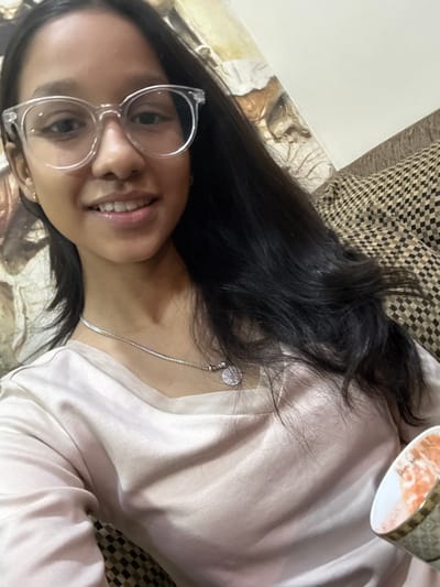 A woman with glasses lies on a patterned surface. She has long, dark hair, a silver necklace, and a light pink satin top. Part of a cup is visible in the top right corner. The woman smiles towards the camera.
