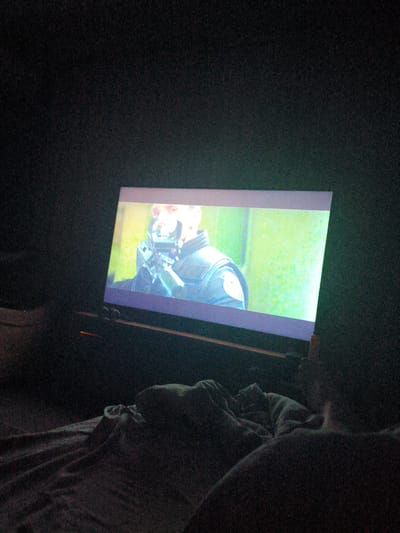 The image shows a person in bed, watching a projection on a screen. The projection displays a close-up of a man in a dark uniform, holding what appears to be a weapon or piece of equipment. The room is dark, with only the light from the projection illuminating the scene. The bedding is dark and appears to be rumpled. There are no visible indications of the specific location within Kurskaya, Russia.