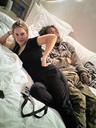 A blonde woman in a black top and bracelet lies in a white bed, gazing at the camera. Beside her is another person, also in black, facing away. A dark-haired individual is nestled amidst blankets and clothing near the headboard. A pair of glasses, a black strap attached to a device, and a small black object rest on the white sheets. A bedside table with items is visible.
