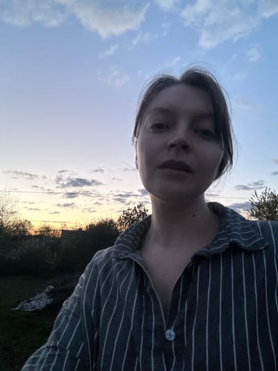 This outdoor selfie, rotated 90 degrees clockwise, captures a person from a low-angle perspective against a natural sky and landscape. The person's face is positioned on the left side of the frame, looking down directly at the camera with a neutral, perhaps contemplative, expression. They have dark hair and are wearing a dark shirt with thin, light-colored horizontal stripes. Part of their upper torso and shoulder are visible on the right, with a single white button visible on the shirt.

The background prominently features a partially cloudy sky, which is light blue with scattered white and grey clouds. A distinct horizon line runs along the right side of the image (which would be the bottom if the image were correctly oriented), showing a line of silhouetted trees and bushes. A warm, golden light emanates from the horizon, strongly suggesting the time of day is late afternoon or early evening, during sunset or dusk. A thin, dark line, possibly a power line or wire, traverses the sky horizontally.

The setting appears to be a natural outdoor environment, likely rural or suburban, consistent with the provided location context of Aksay, Kazakhstan. The overall lighting is soft and subdued. No visible text is present in the media file.