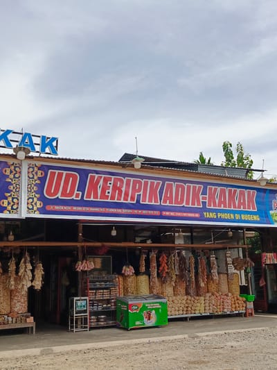 The image shows a storefront in Sawang, Indonesia, featuring a banner with the text "UD. KERIPIK ADIK-KAKAK" in large red letters. Below this, smaller text indicates the location as "JL. BANUA ACEH - MEDAN DESA SAWANG KEC. PEUSAADAH KAB. BIREUEN". The store appears to sell various snacks, with many bags and packages displayed both inside and hanging outside. A green freezer chest, labeled "Azizah Ice Cream", is visible to the right of the entrance, and a shelf unit with more products is located inside. The sky is overcast, and the ground in front of the store is unpaved and appears to be gravel. The building has a corrugated metal roof, and several light fixtures are visible. There are no people clearly visible in the image.