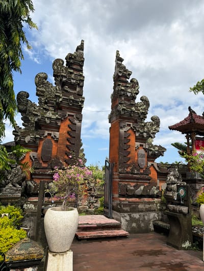 This is a wide-angle, eye-level, outdoor, daytime photograph of the Candi Bentar gate at the Goa Lawah Temple in Klungkung, Indonesia. The gate, made of red brick and dark stone with mossy growth, is split into two towers, with a smaller, ornate pavilion visible to the right. In the foreground, a large, light-colored pot with a blooming pink bougainvillea plant sits on a pedestal, and to the left, lush green foliage hangs from a tree. A stone fountain with a sink is present to the right of the gate, and a paved courtyard with scattered puddles reflects the cloudy sky. No people are present in the image.