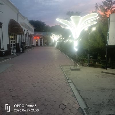 The image depicts a paved walkway leading towards a building at dusk. The walkway is made of interlocking reddish-brown pavers and is bordered by concrete on the right. Several large, illuminated decorative structures resembling stylized flowers or petals are positioned along the right side of the walkway, casting a bright white light. In the background, a white building with a dark trim and arched windows is visible, with some neon signage illuminated in red above one of the entrances. Trees and foliage are present behind the decorative lights and to the left of the building. The sky is a soft gradient of grey and pink, indicating twilight. The timestamp in the lower left corner indicates the photo was taken on June 27, 2026, at 21:38. This scene is located in the city of Atyrau, Kazakhstan, likely in front of a restaurant or entertainment venue given the decorative lighting and signage.