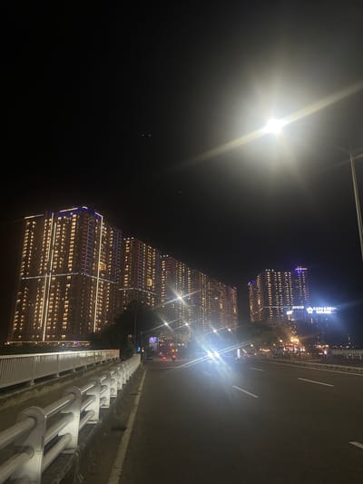 The image depicts a nighttime scene of a city skyline and a road, likely taken from an elevated perspective such as a bridge or overpass. The foreground features a road with distinct white lane markings, adjacent to a barrier system composed of parallel white pipes and a metal grating. In the background, several tall buildings are illuminated with a multitude of lights, creating a vibrant urban landscape against the dark night sky. The buildings exhibit a modern architectural style, with many windows reflecting the internal lighting. Some buildings are adorned with decorative colored lights, particularly along their edges, with prominent blue and red lighting visible.
A bright, powerful light source, possibly a streetlamp or vehicle headlight, is prominently visible in the upper left quadrant, casting a strong glare and creating lens flare effects that obscure details in that area. The overall ambiance is one of a bustling city at night, with the illuminated architecture suggesting a commercial or residential district.
The visible text on one of the buildings reads "CASARENE GOLDE." The image conveys a sense of urban energy and nighttime activity. The time of day is clearly night, and the absence of visible weather elements suggests clear conditions. The location cues are the modern, illuminated skyscrapers and the road infrastructure, consistent with an urban environment.